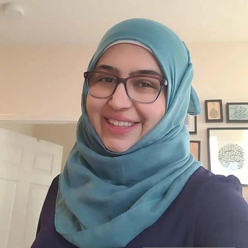 Smiling person wearing a blue hijab and glasses, with framed artworks in the background.